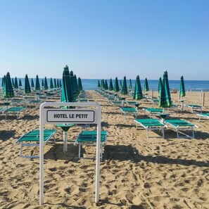 Ein Strand mit Reihen aus grünen Sonnenschirmen und Liegestühlen, ein Schild mit der Aufschrift 'HOTEL LE PETIT'.