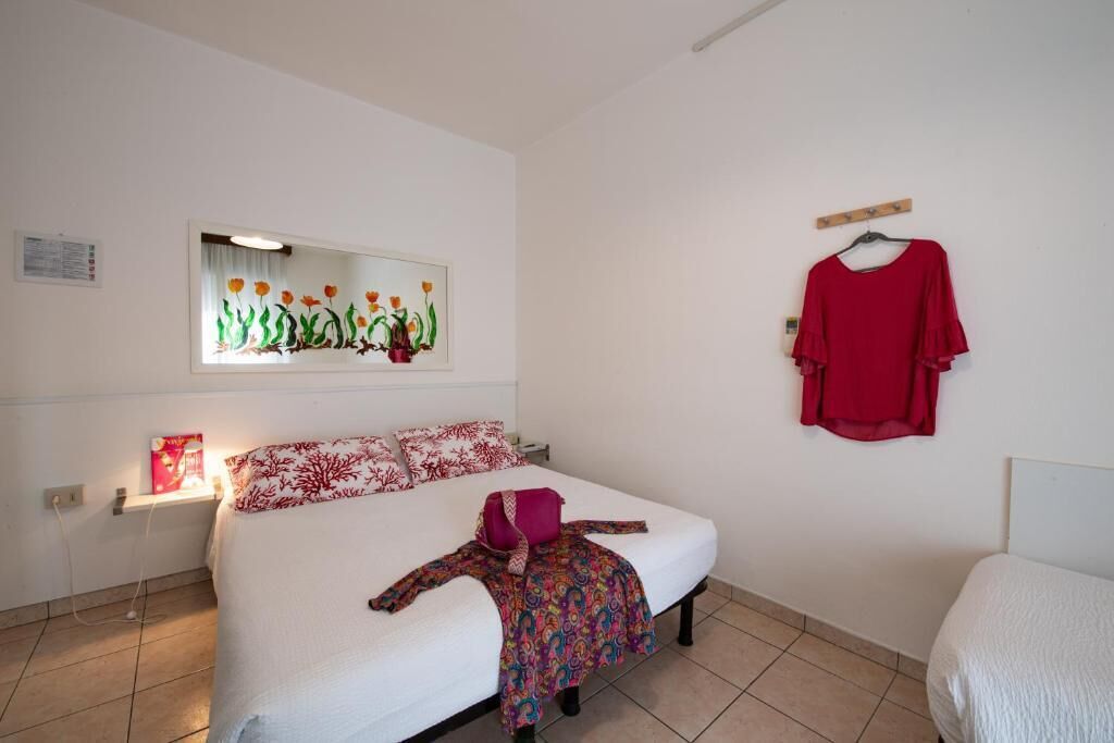 A bedroom with a bed, a mirror, a red shirt on a hanger, and a window with a view of flowers.