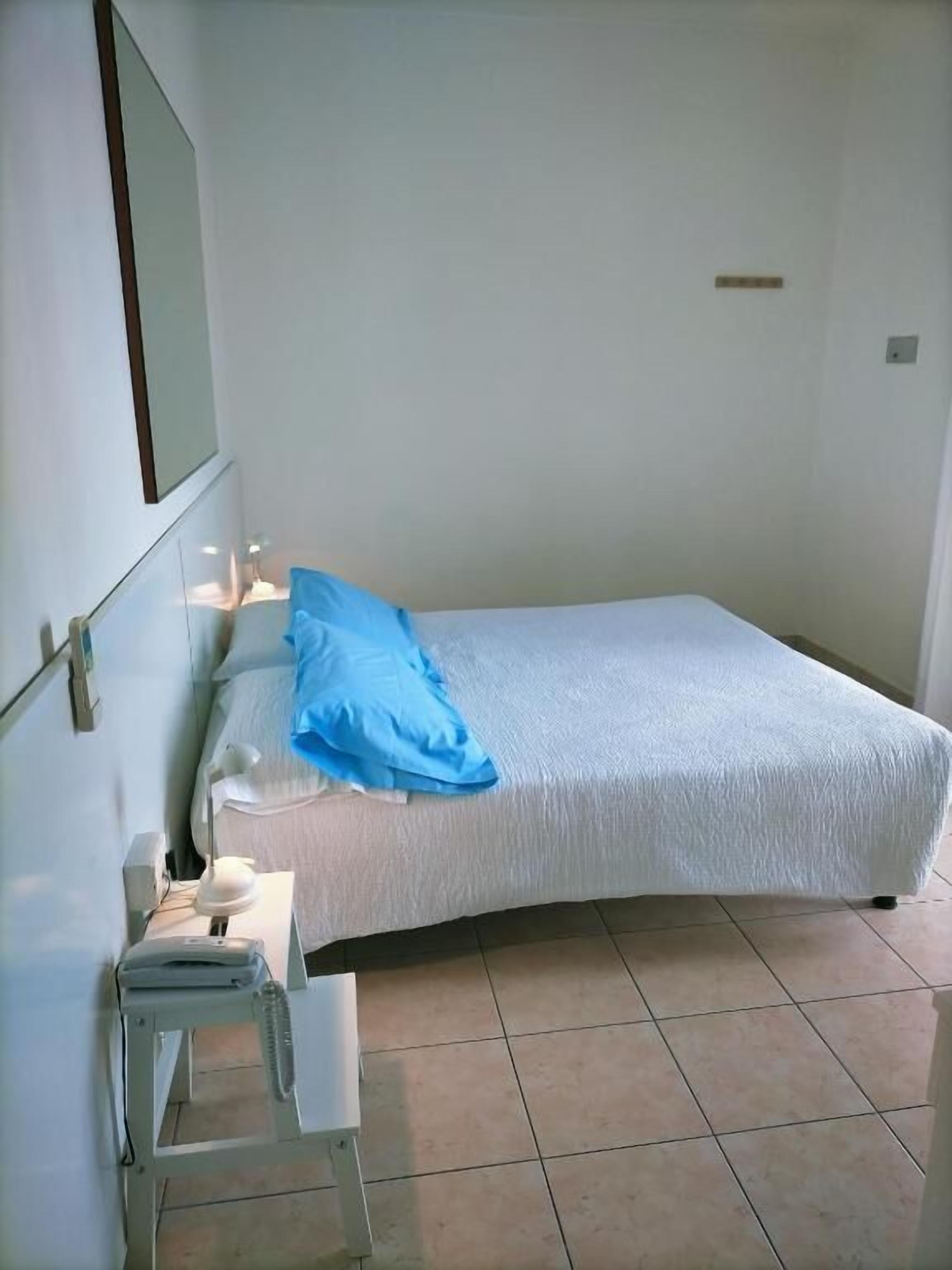 A single-bed room with a white bedspread, a blue pillow, a bedside table with a lamp, and a small white chair.