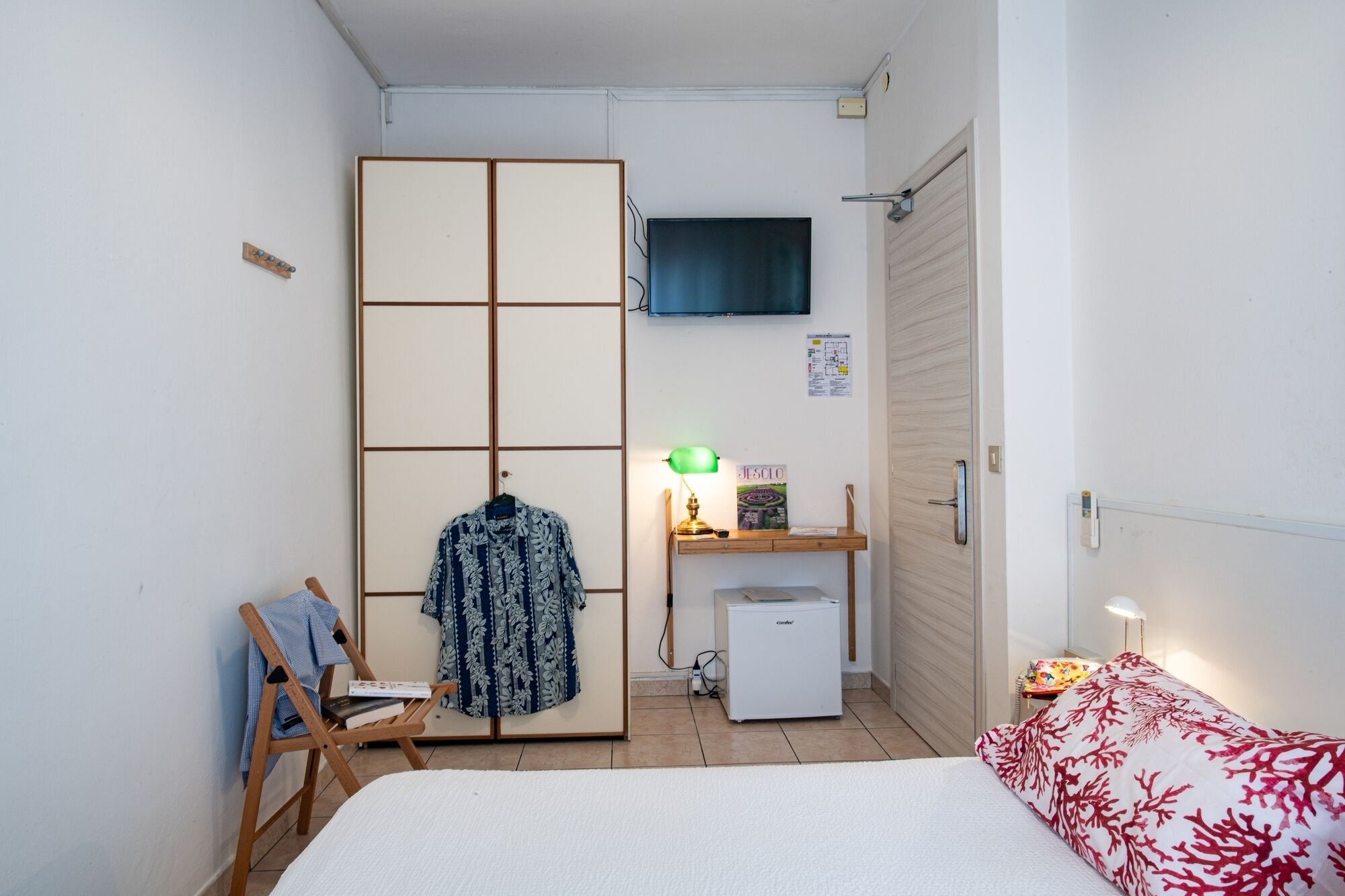 A compact bedroom with a bed, a wardrobe, a desk with a lamp, a small fridge, and a TV mounted on the wall.