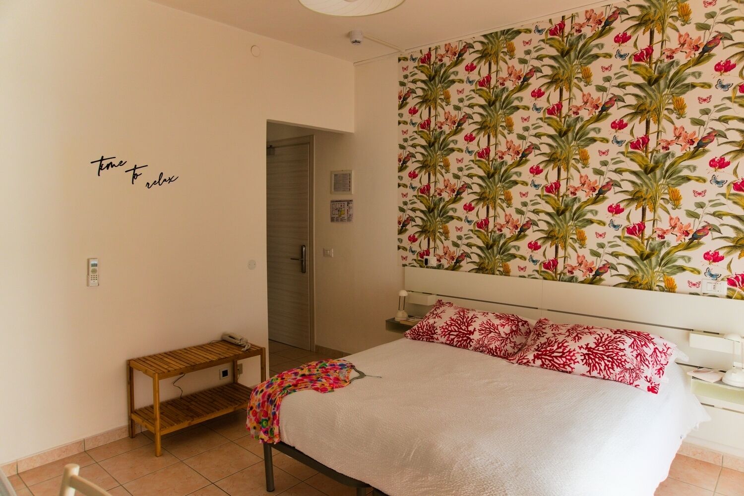 A bedroom with a bed, a small wooden bench, a door, and a wall with a floral pattern.