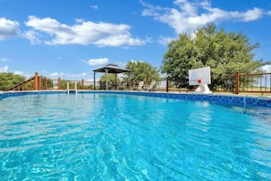 Una piscina azul cristalina con un deck cercado, una zona de estar cubierta y árboles al fondo.
