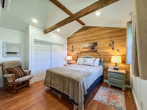 A bedroom with a wooden ceiling, a bed with a headboard, a nightstand, a chair, and a door. - Wagon Wheel Cottage Couples Getaway in the Country (Waco)