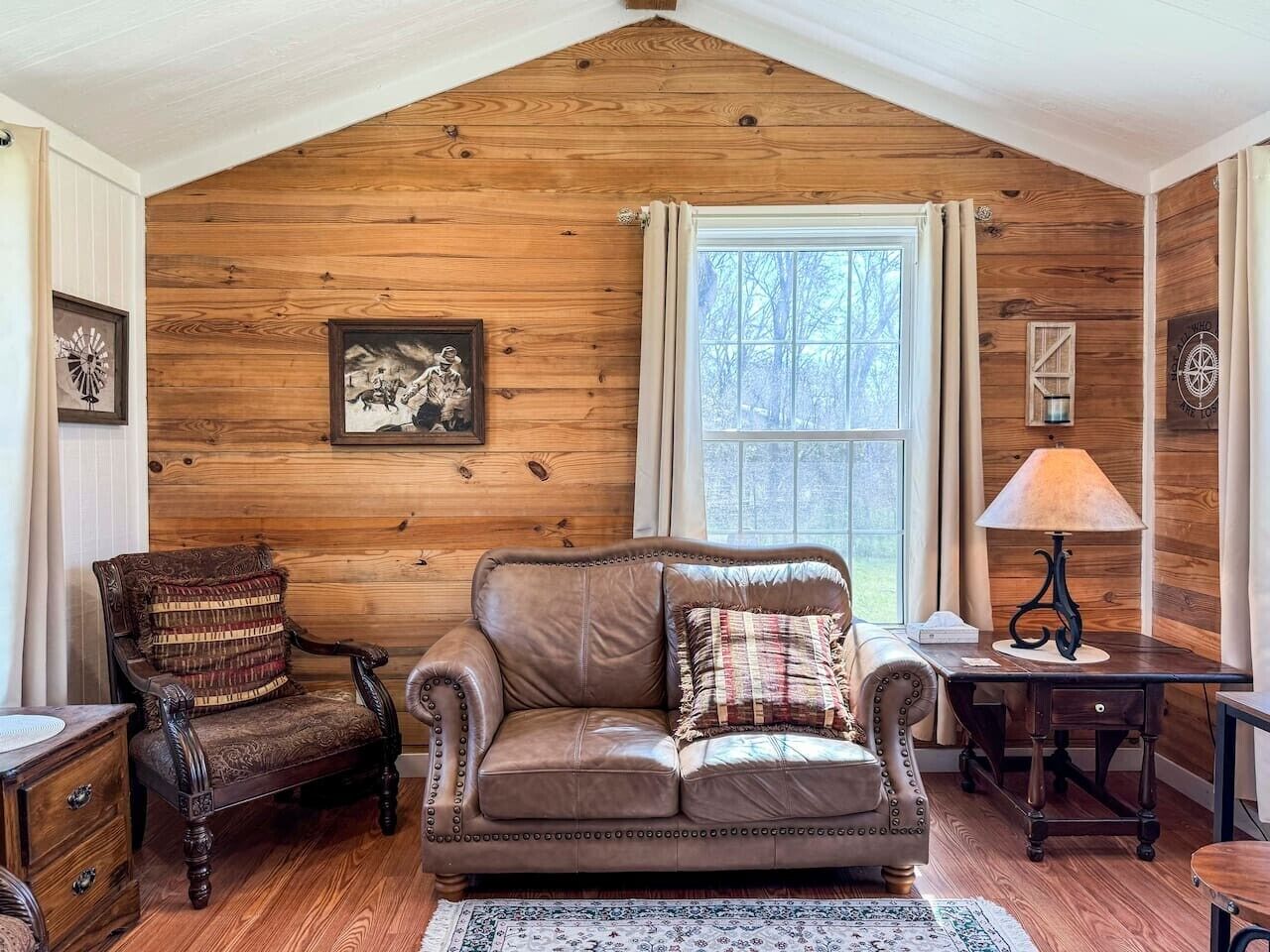 A cozy room with wooden walls, a leather sofa, a wooden chair, a table with a lamp, and a window with curtains.