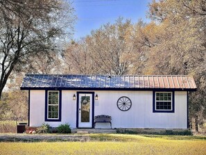 A small, single-story building with a white exterior and dark trim, a front door, two windows, a wheel-like decoration, and a bench outside. - Wagon Wheel Cottage Couples Getaway in the Country (Waco)