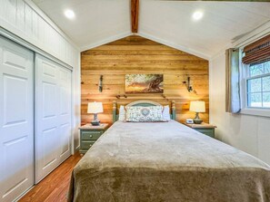 A bedroom with a wooden ceiling, a bed with a patterned headboard, two bedside tables with lamps, and a window with curtains. - Wagon Wheel Cottage Couples Getaway in the Country (Waco)