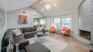 A spacious living room with a gray sectional sofa, a glass coffee table, and a fireplace. There is a Buddha painting on the wall and a view of the outside through large windows. - Your Houston Vacation Getaway, 4-BD, Pool, Patio (Houston)