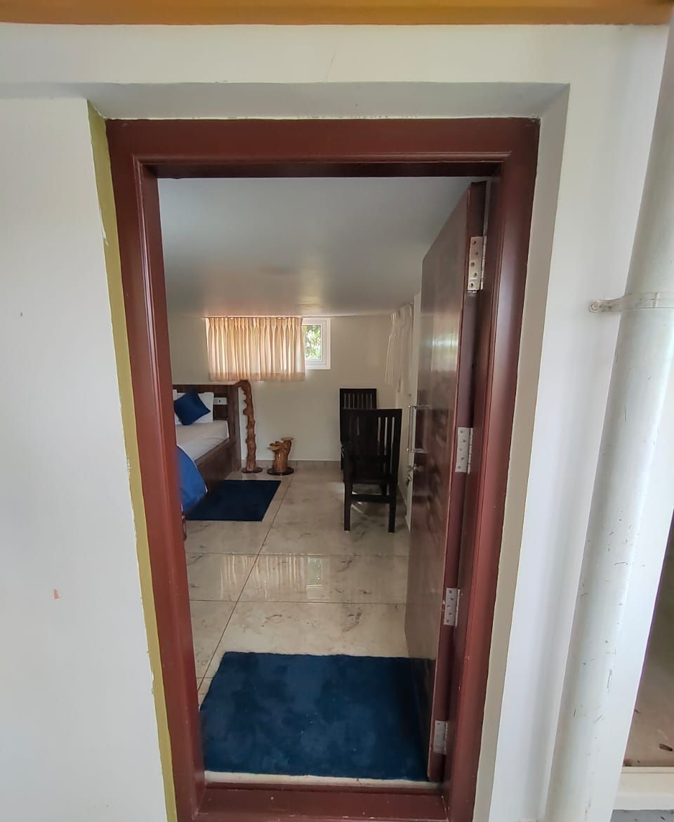 A doorway leading to a room with a bed, a chair, a window with curtains, and a small table.