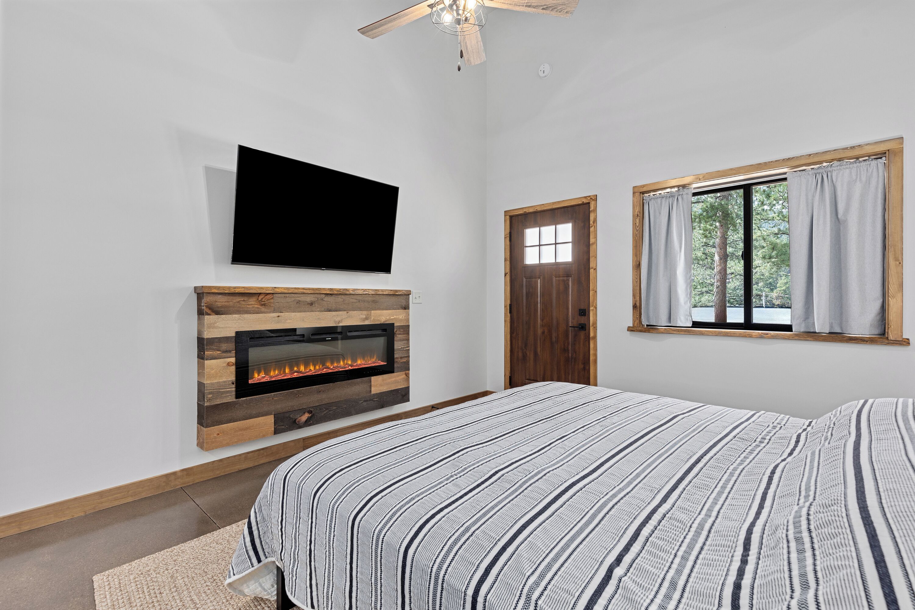A bedroom with a fireplace, a TV mounted on the wall, a window with curtains, and a bed with striped bedding.