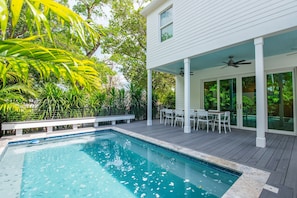 Une piscine avec une terrasse en bois, un patio couvert avec des chaises et un bâtiment blanc doté de grandes fenêtres.