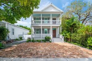 A two-story house with a front porch, a brick driveway, and surrounding greenery. - The Rigby House - Pool, One block from Duval St (Key West)