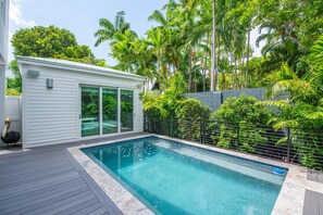 A swimming pool with a wooden deck, surrounded by lush greenery and a white building with glass doors. - The Rigby House - Pool, One block from Duval St (Key West)