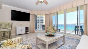 A spacious living room with a flat-screen TV, a ceiling fan, a large window with a view of the beach, a sofa, a coffee table, and a cabinet. - Serene Beachfront Condo w/ Beach Service in Central Destin! 🌴🐚🌊 (Destin)