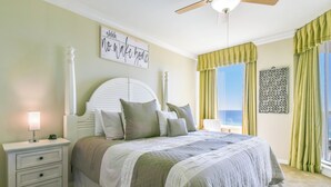 A bedroom with a bed, nightstands, a ceiling fan, a window with curtains, and a wall decoration. - Serene Beachfront Condo w/ Beach Service in Central Destin! 🌴🐚🌊 (Destin)