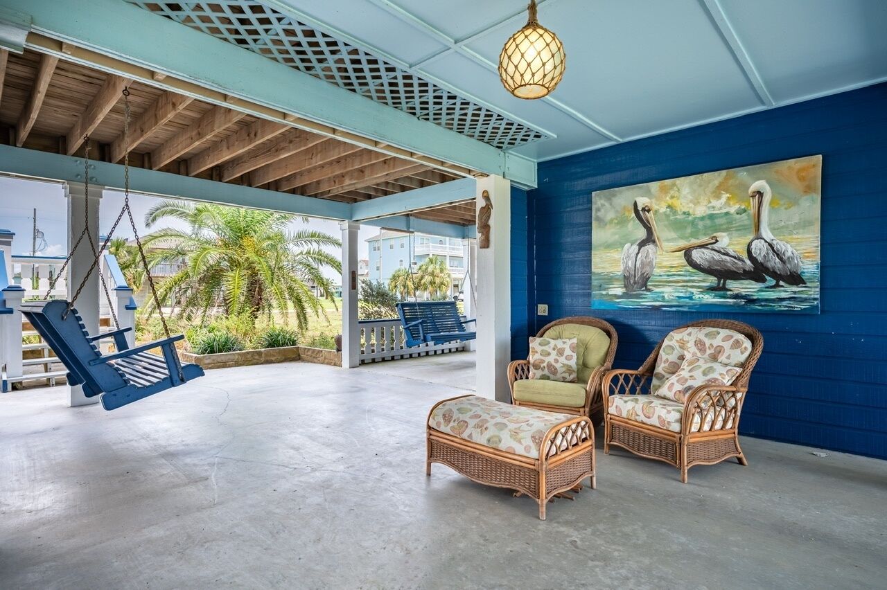 A covered outdoor area with wicker furniture, a swing chair, and a painting of pelicans.