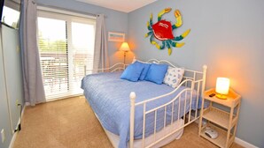 A bedroom with a bed, a bedside table with a lamp, a wall decoration, and a window with curtains. - Sunset Landing 24 2 Bedroom Townhouse by RedAwning (Ocean City)