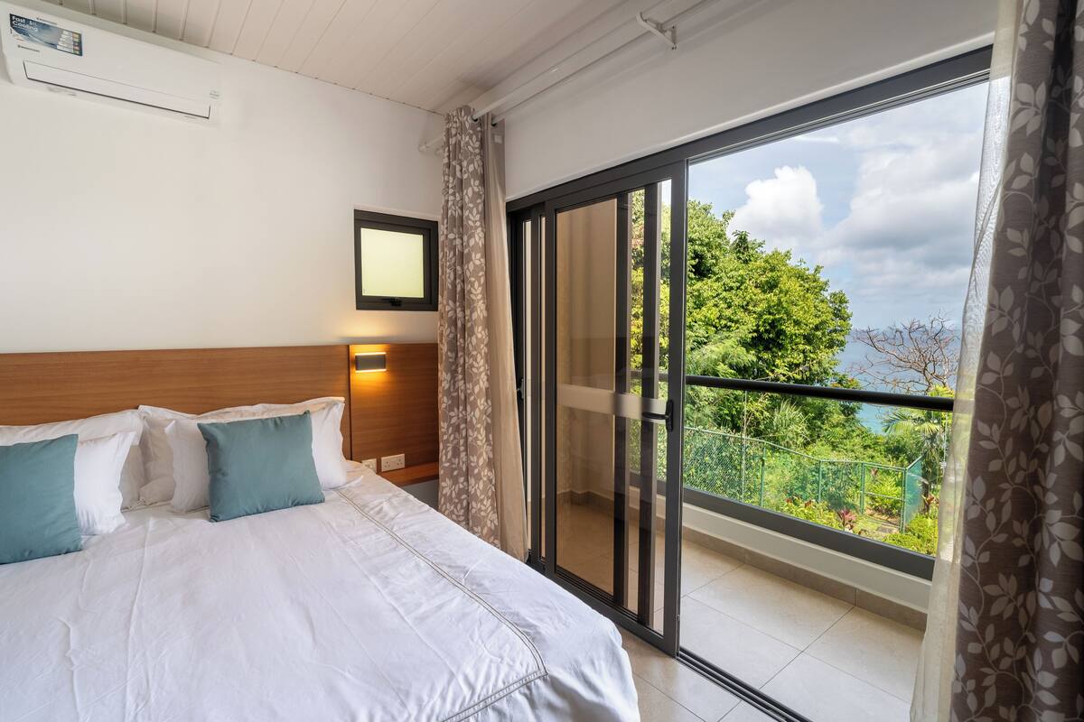A hotel room with a bed, a nightstand, a window with a view of greenery, and a sliding glass door to the balcony.