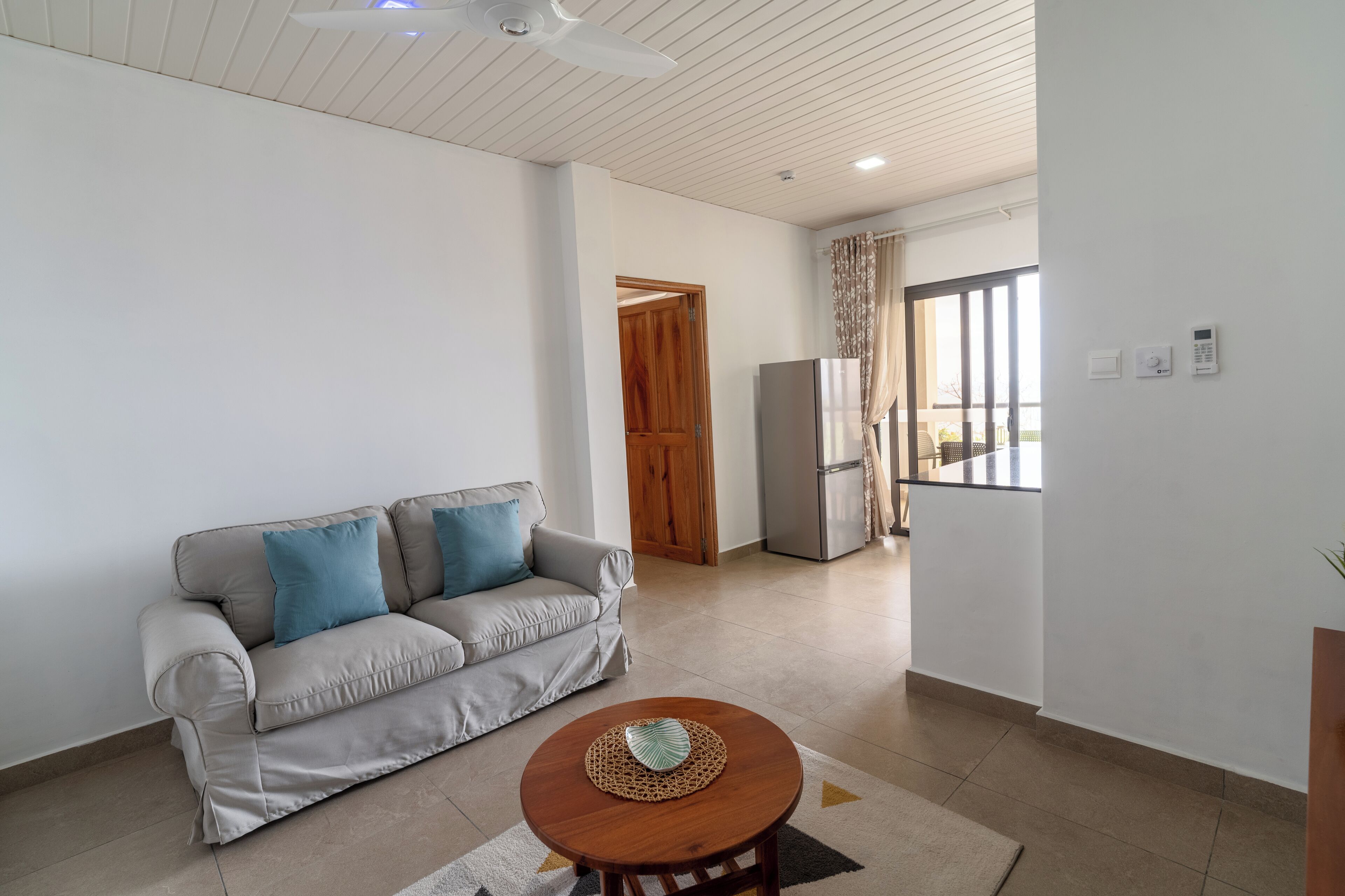 A living room with a sofa, a coffee table, a ceiling fan, a refrigerator, and a sliding glass door leading to a balcony.