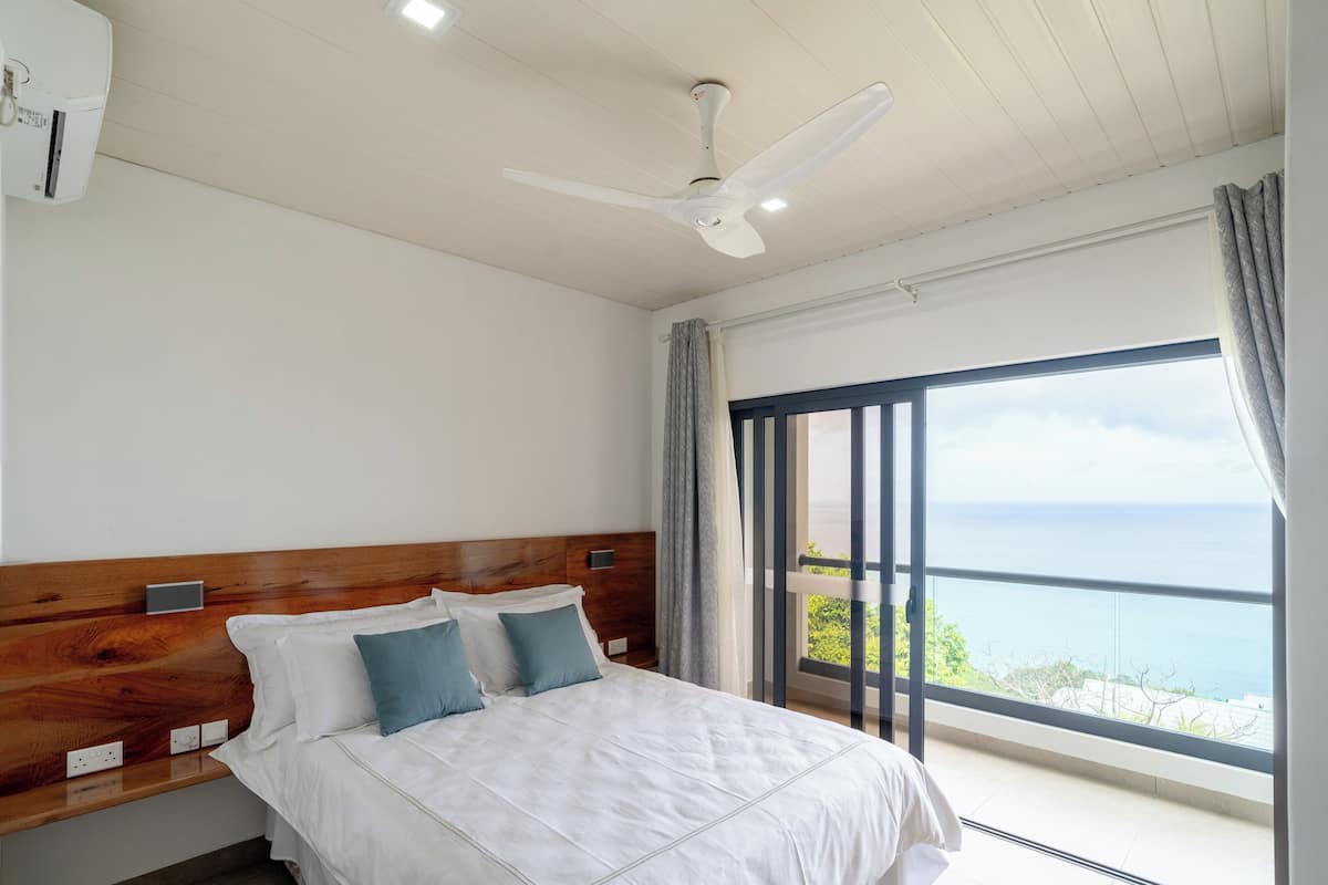 A bedroom with a large bed, a ceiling fan, a window with a view of the sea, and a wooden headboard.