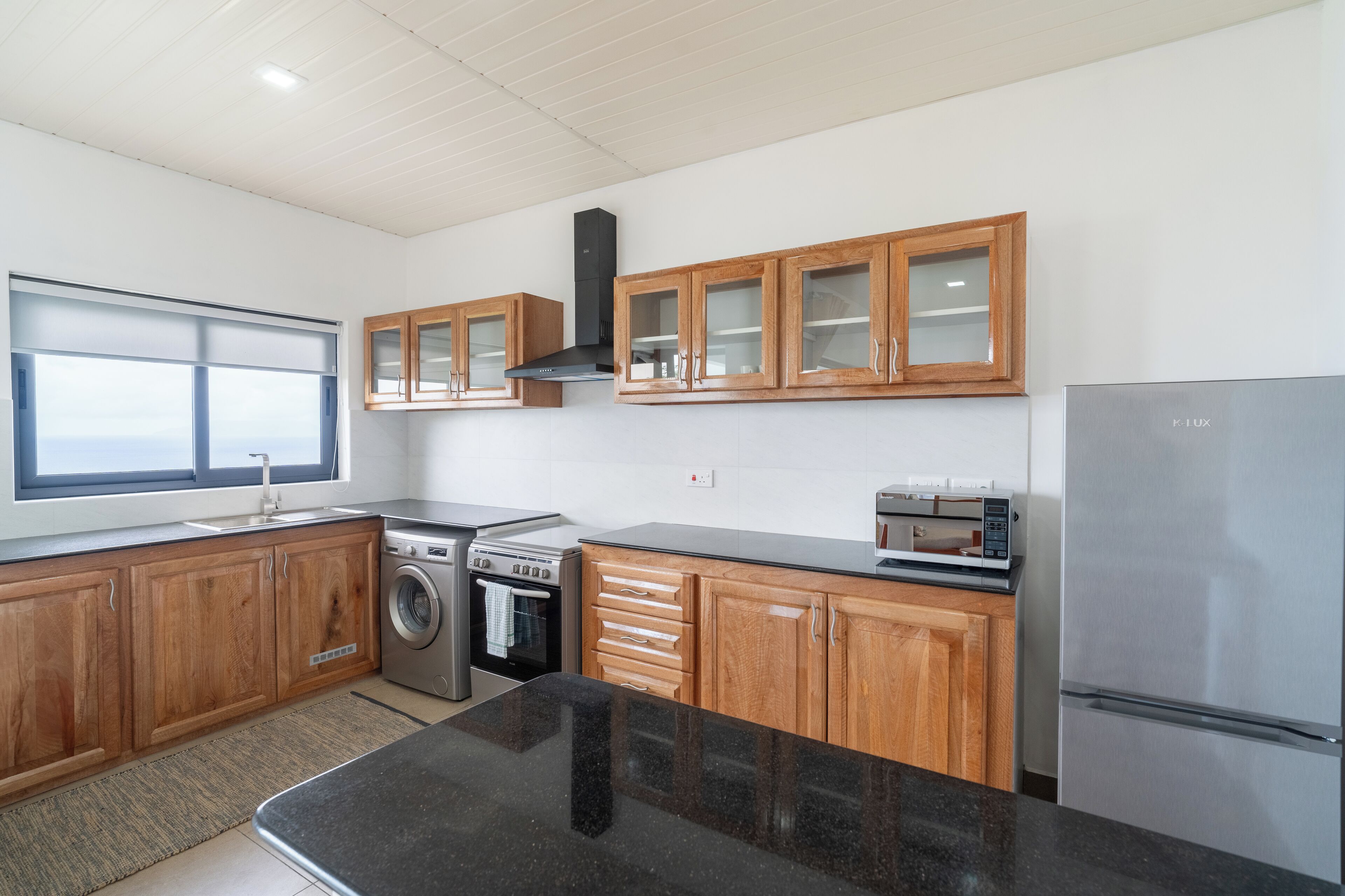 A modern kitchen with wooden cabinets, a stainless steel refrigerator, a washing machine, and a microwave.