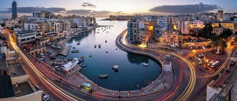 Un paisaje urbano costero con una glorieta, un puerto deportivo y edificios a lo largo del litoral.