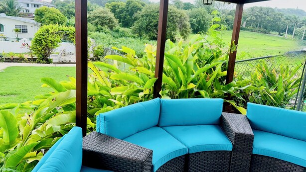 A covered outdoor seating area with a curved blue sofa and wicker furniture, surrounded by lush greenery and a view of a residential area.