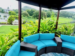 Un espacio al aire libre techado con un sofá azul curvo y mobiliario de mimbre, rodeado de abundante vegetación y vistas a una zona residencial.