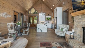 A spacious interior with wooden walls, a dining area with white chairs, a kitchen with white cabinets, and a stone fireplace. - Elevated Escape Blue Ridge GA (Blue Ridge)