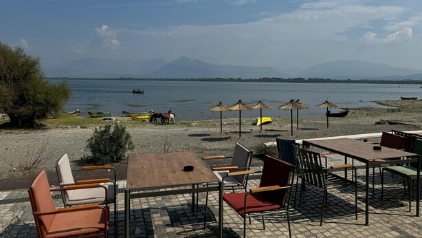 An outdoor seating area with tables and chairs, a beach with boats, and a mountain in the background. - Hotel Flaer (Shkodër)
