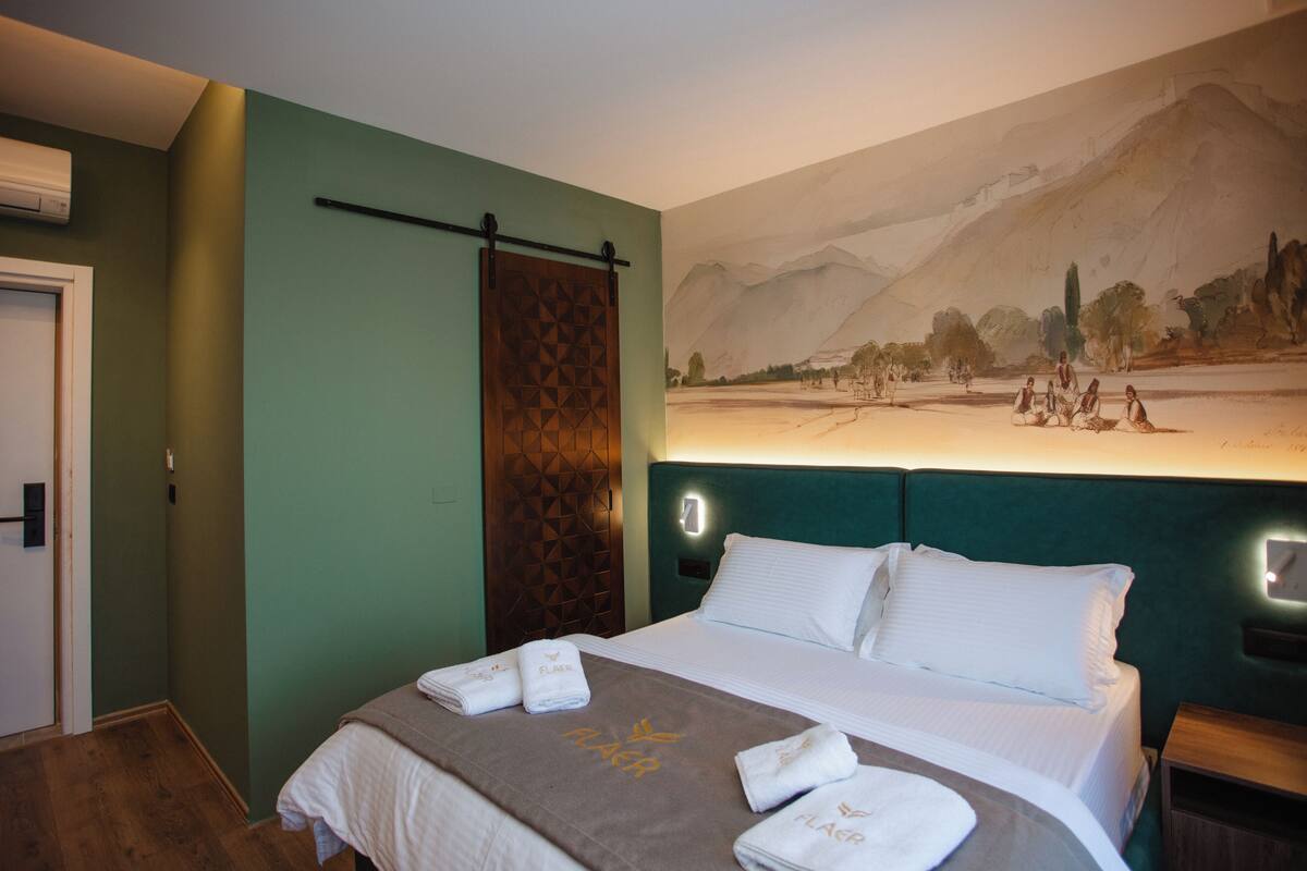 A hotel room with a bed, a wall-mounted artwork depicting a landscape, and a door.