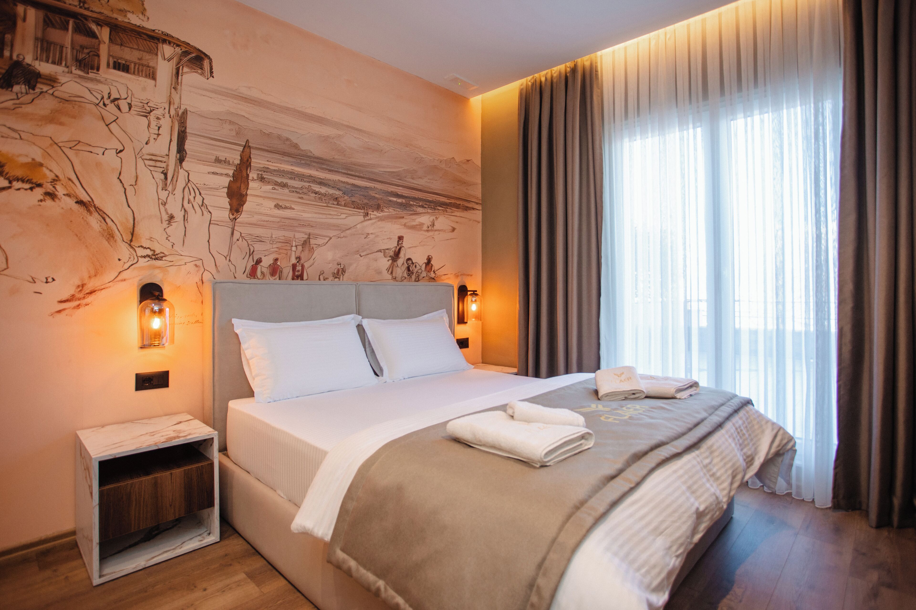 Una habitación de hotel moderna con una cama grande, una mesita de noche, un mural en la pared y un ventanal con cortinas.