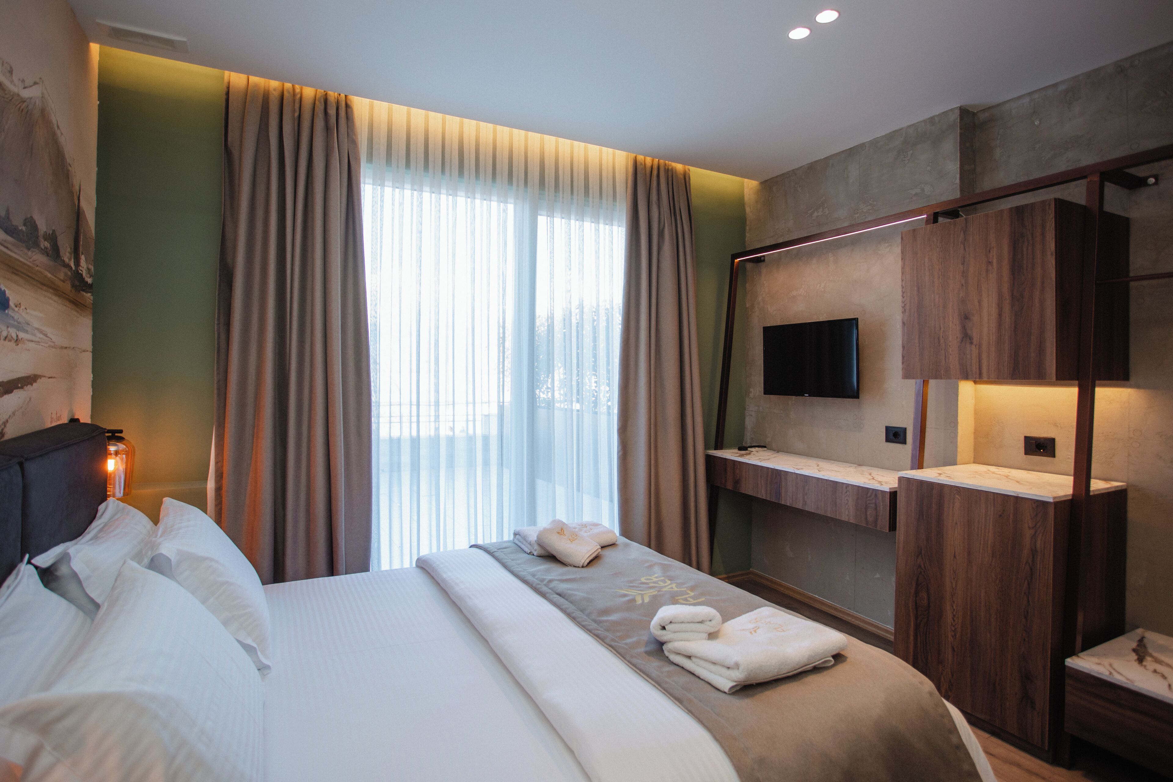 Une chambre d’hôtel moderne dotée d’un grand lit, d’un téléviseur à écran plat fixé au mur, d’une coiffeuse en bois et d’une grande fenêtre avec des rideaux.