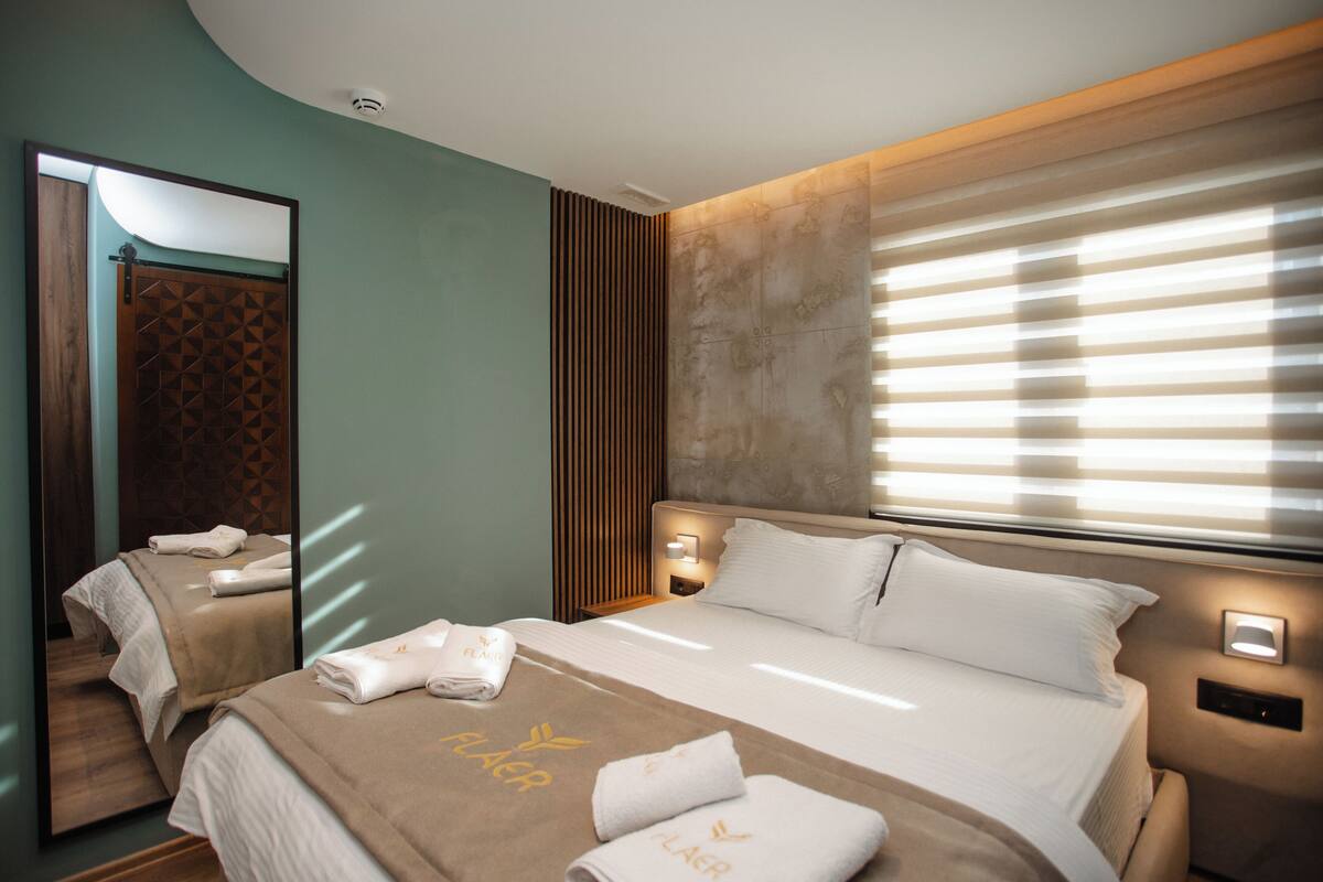 A hotel room with a large bed, a mirror, a nightstand, and a window with blinds.