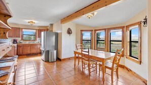 A spacious kitchen with wooden cabinets, a stainless steel refrigerator, and a dining area with a wooden table and chairs. Large windows provide views of the outdoors. - Peggy s View - Uplifting Cozy Mountain Views (Alpine)