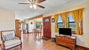 A spacious living room with a ceiling fan, a wooden TV stand, and a view through large windows. - Peggy s View - Uplifting Cozy Mountain Views (Alpine)