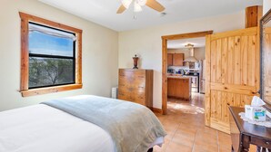 A bedroom with a bed, a ceiling fan, a wooden dresser, and a view of the kitchen through an open door. - Peggy s View - Uplifting Cozy Mountain Views (Alpine)