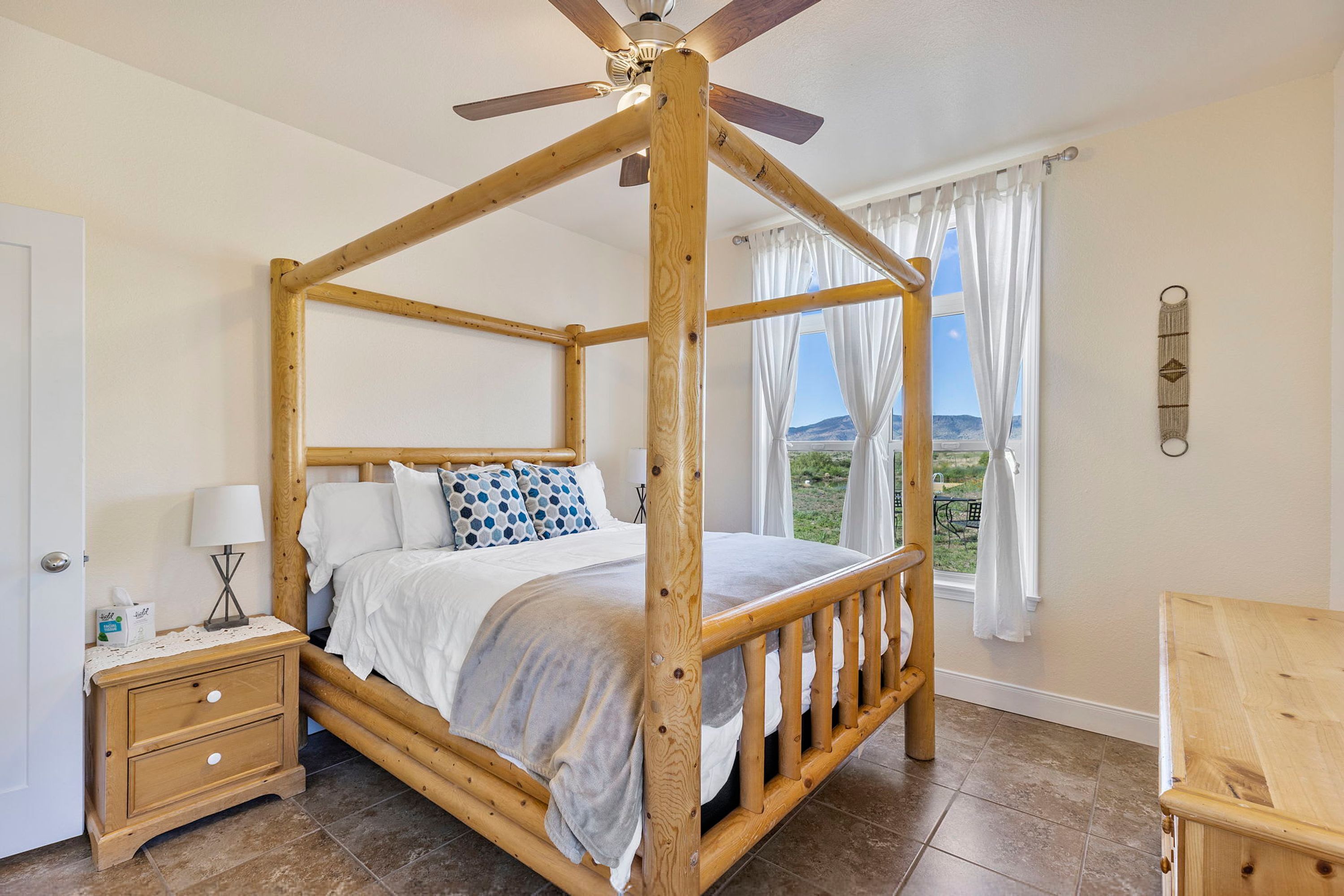 Una cama con dosel de madera, ventilador de techo, ropa de cama blanca y vistas al exterior a través de una ventana con cortinas blancas.