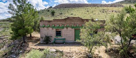 Un pequeño edificio de ladrillo con una puerta y ventana verdes, rodeado de vegetación escasa y una colina al fondo.