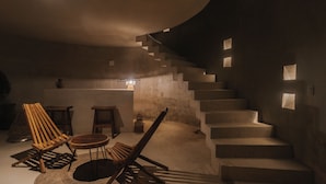 A dimly lit room with a curved staircase, wooden chairs, a small table, and a rug. - Seremonia Selva (Tulum)