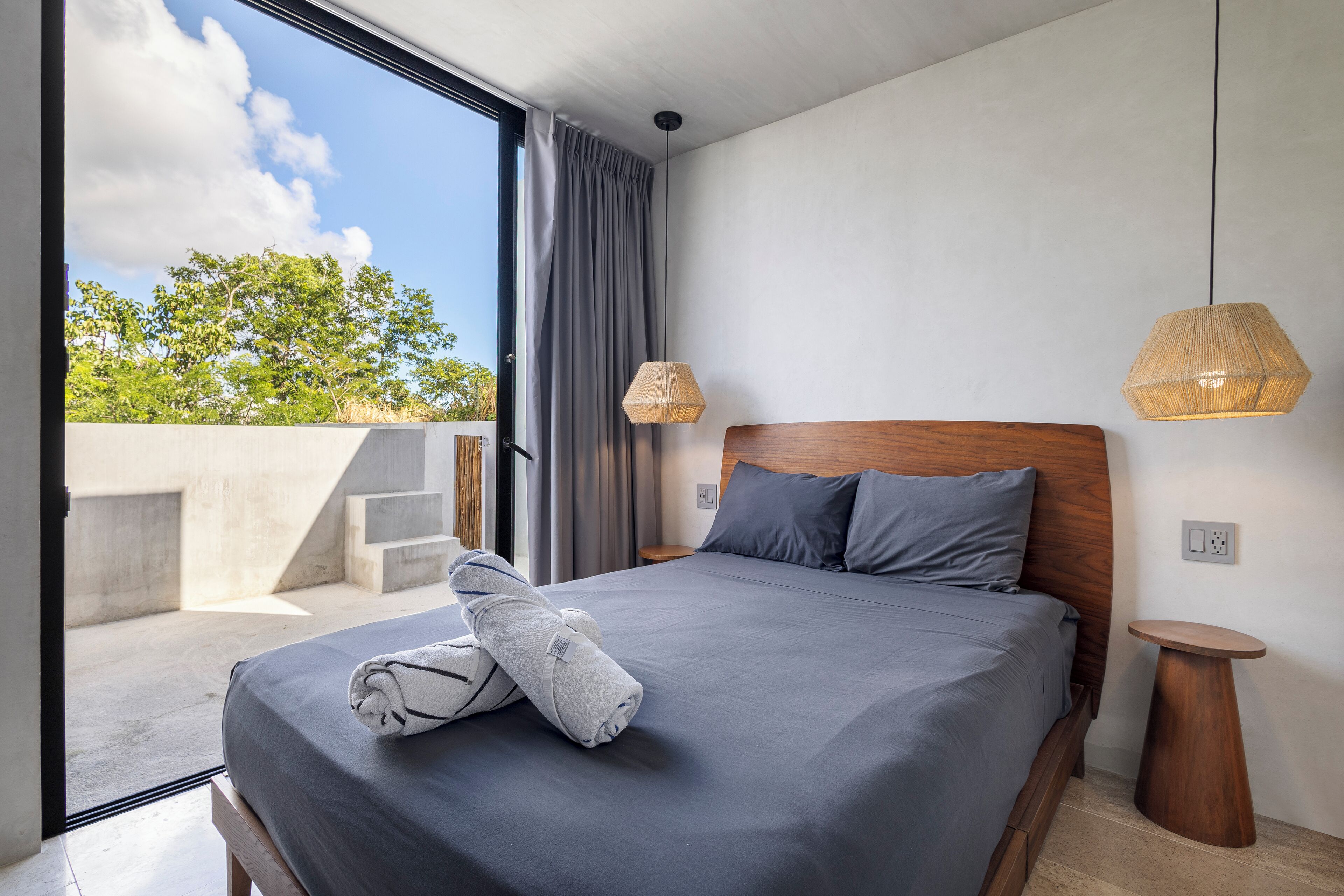 A modern bedroom with a large bed, grey bedding, and a view of greenery outside.
