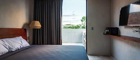 Un dormitorio con cama, televisor, una pequeña estantería, ventana con cortinas y vistas al exterior.