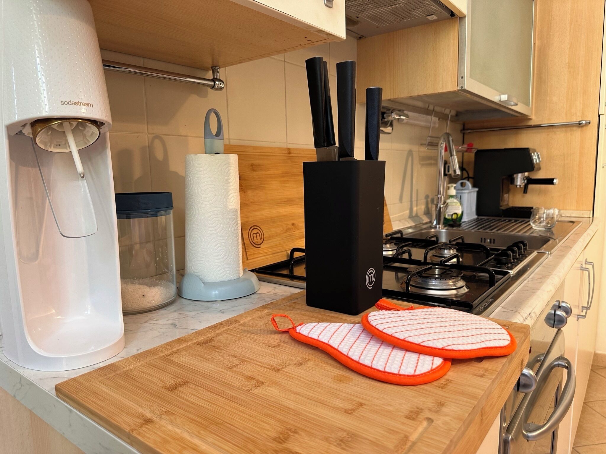 A kitchen with a gas stove, a paper towel dispenser, a cutting board, and a kettle.