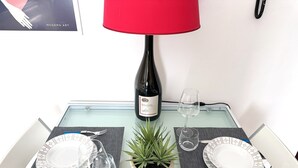 Ein Esstisch ist gedeckt mit Tellern, Besteck, Weingläsern und einer Weinflasche. In der Mitte steht eine rote Lampe und ein Blumentopf.