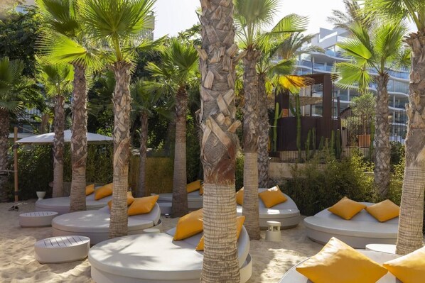 Outdoor seating areas with round white cushions and yellow pillows, surrounded by palm trees and a sandy ground.