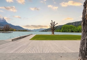 A wooden deck with a grassy area, a tree sculpture, and a lake with mountains in the background. - Casa-Isabelle pres du lac (Embrun)