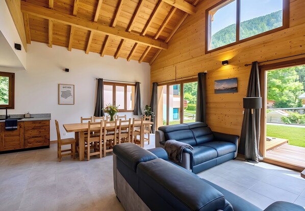 A spacious living area with a wooden ceiling, a black leather sofa, a dining table with chairs, and large windows offering a view of the outdoors.
