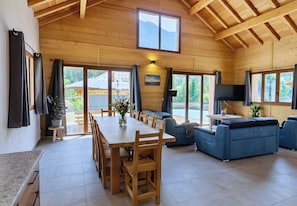 A spacious room with a wooden ceiling, a dining area with a table and chairs, a blue sofa, and large windows offering a view of the outdoors.