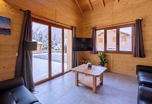 Una habitación acogedora con sofá, mesa, televisión y ventanas amplias que ofrecen vistas a árboles nevados y montañas.