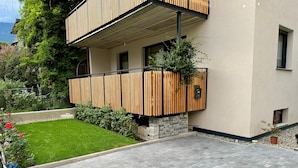 A modern two-story building with a flat roof, a small garden area, and a paved driveway. - Modern apartment with large balcony. Near city center. (Meran)