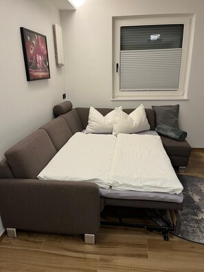 Ein kompakter Wohnraum mit Schlafsofa, einem kleinen Beistelltisch und einem Fenster mit Jalousien.